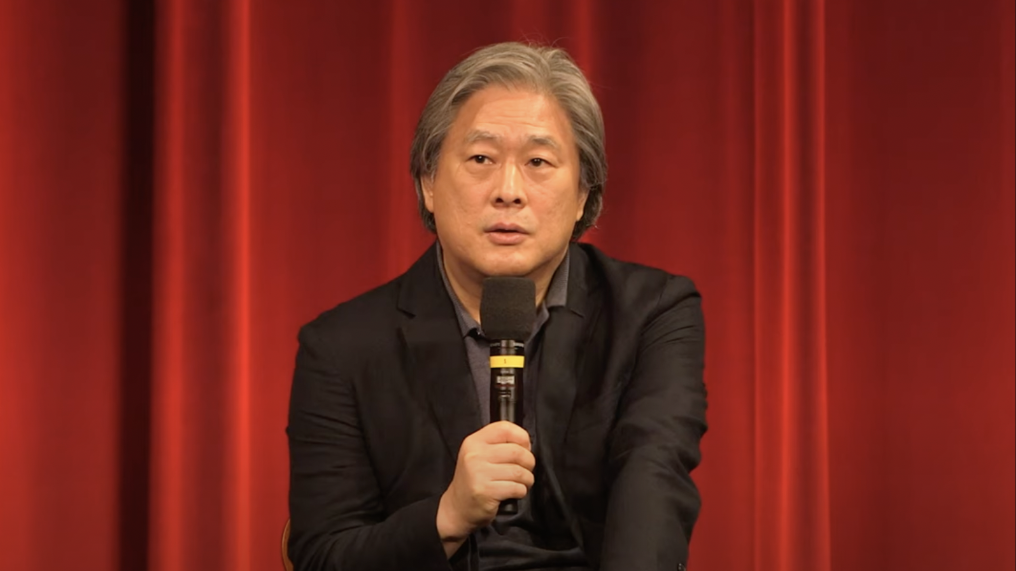 Park Chan-wook speaks into a microphone in front of a red curtain.